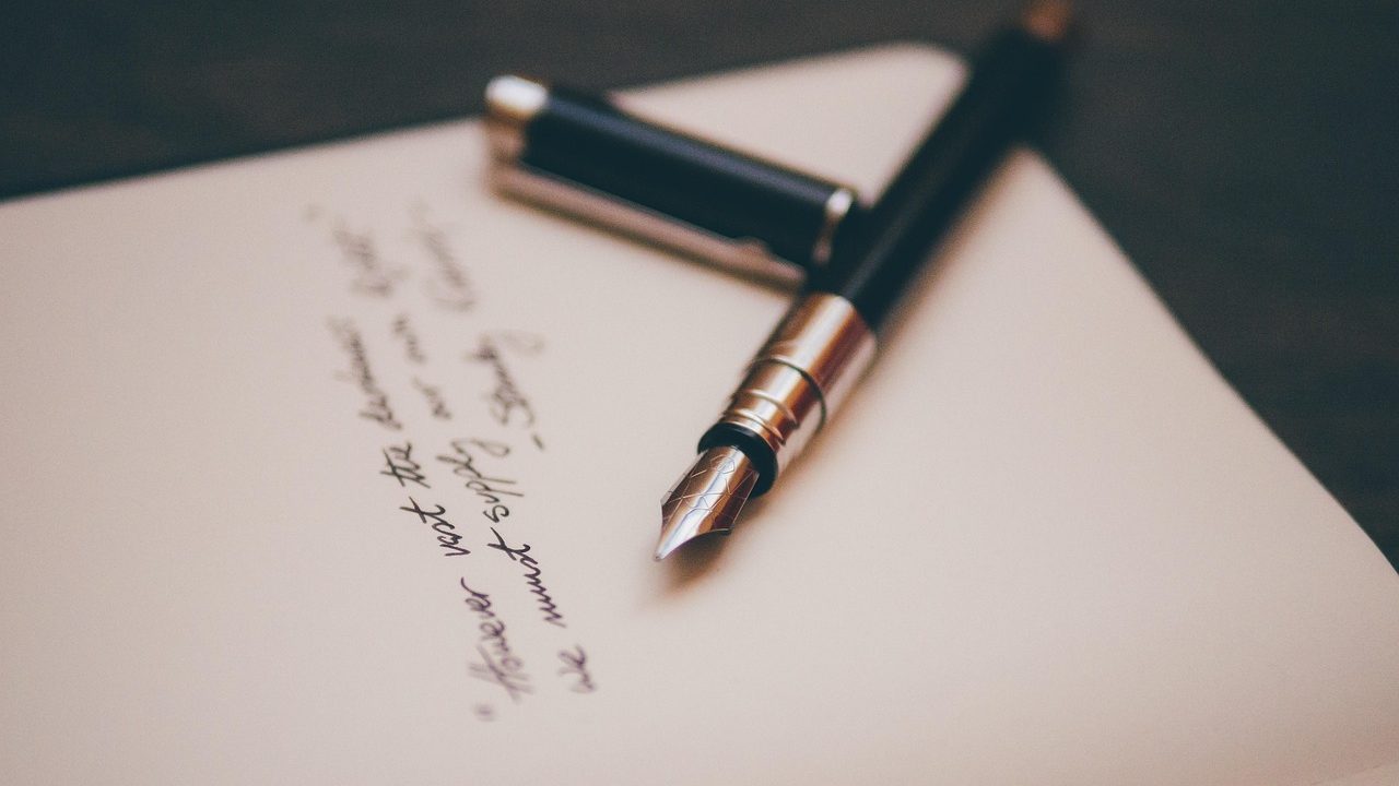 A handwritten letter on cream paper with a fountain pen resting beside it on a wooden desk.