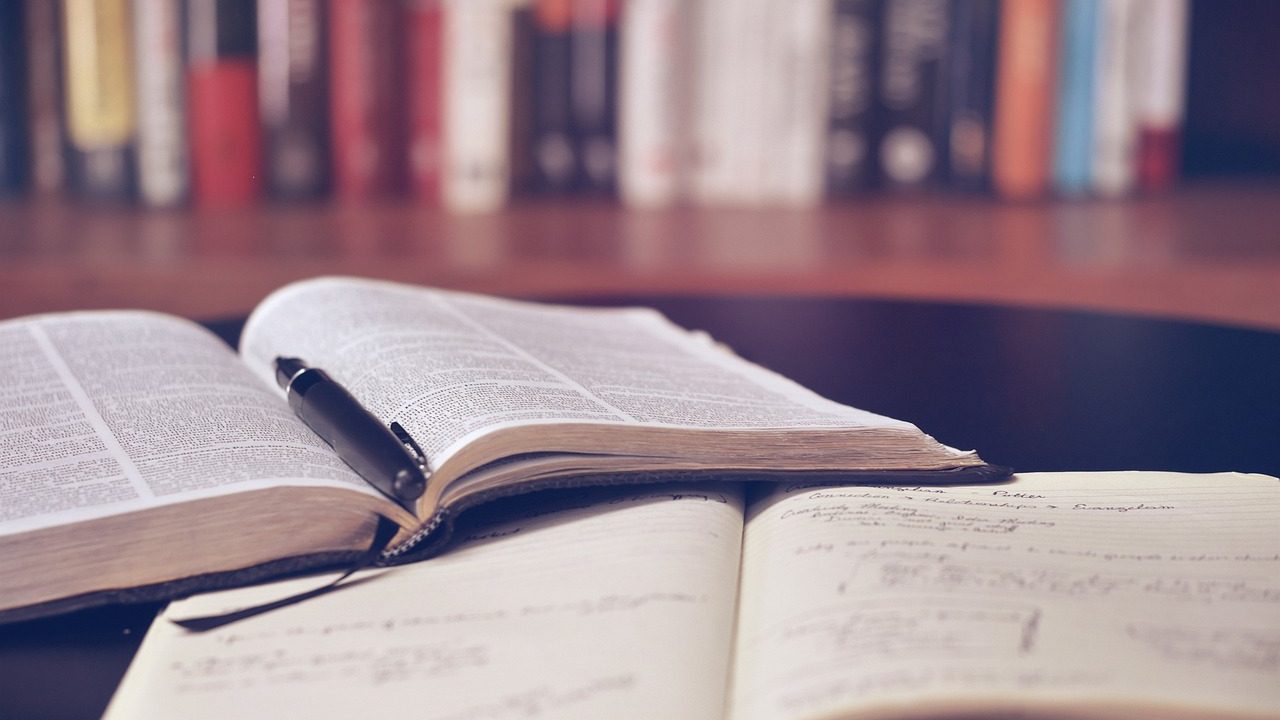 A hardcover book lies open on a wooden table in front of tall library shelves.