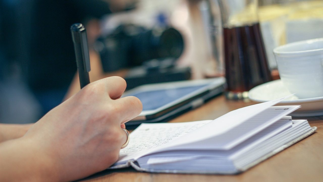 A pen rests on an open notebook beside a stack of books on a wooden desk.