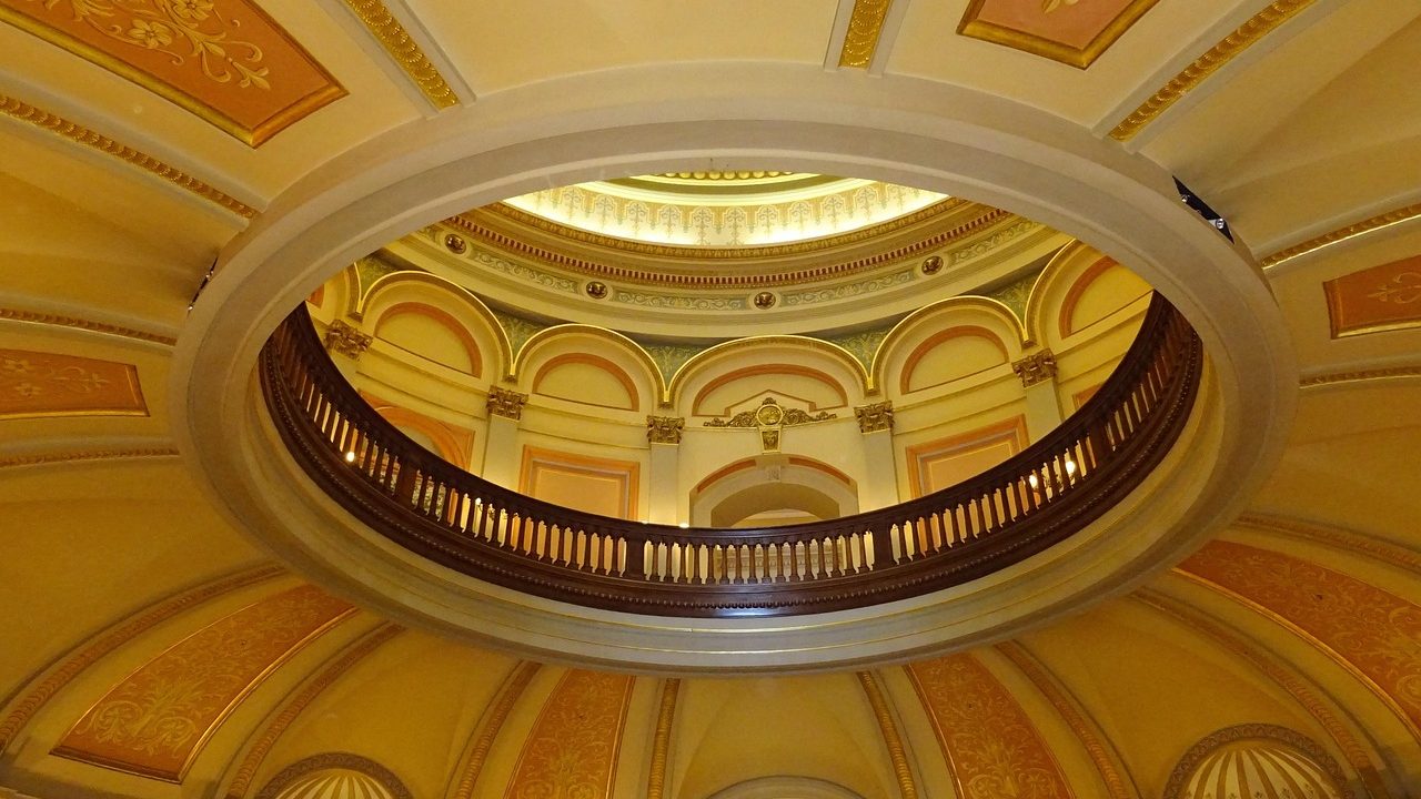 Empty senate chamber with rows of wooden desks under a domed ceiling.