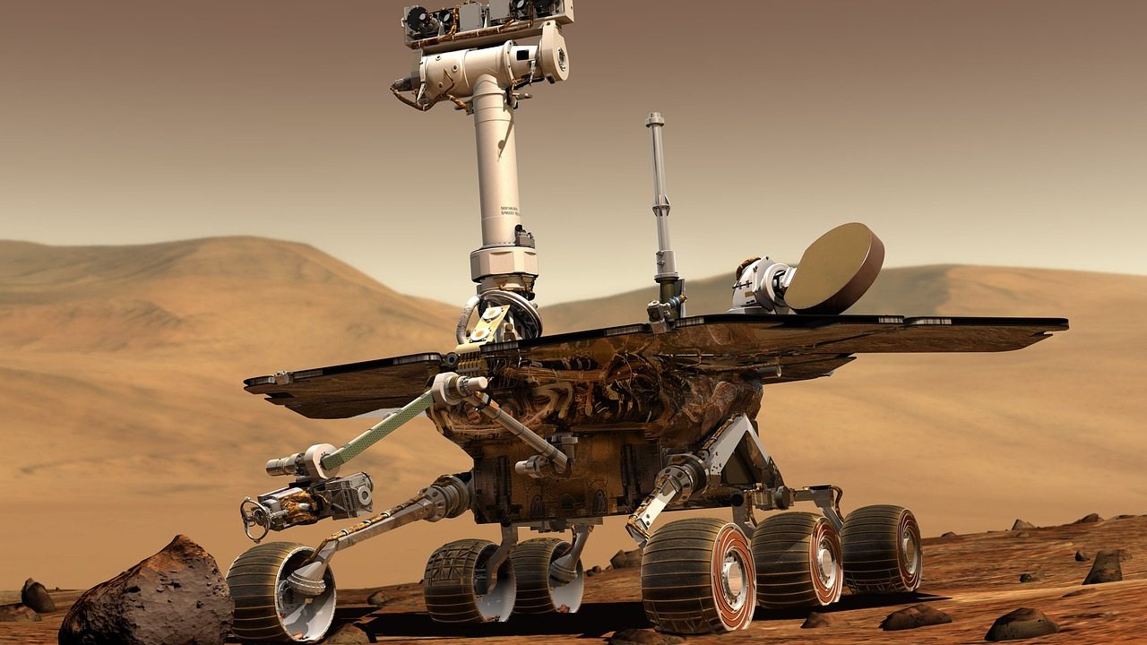 A six-wheeled robotic rover traverses a reddish rocky desert landscape under a pale sky.