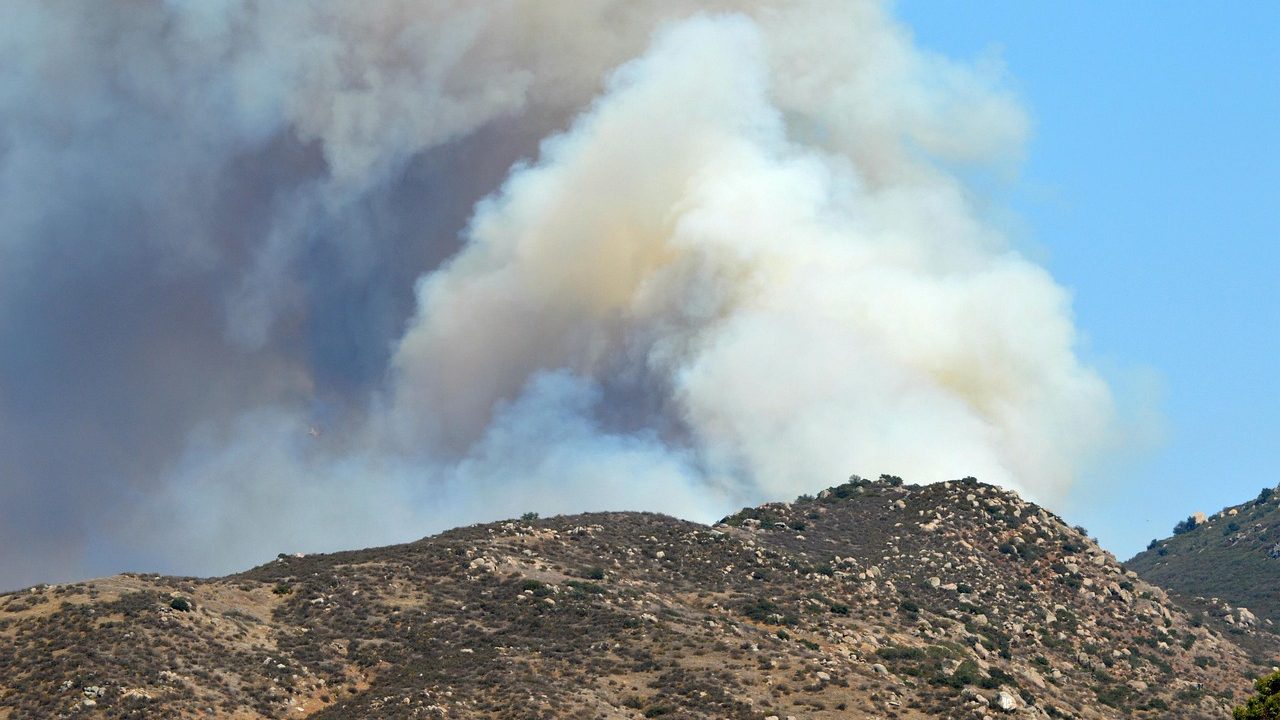 Thick orange smoke rises above a dry hillside as flames advance through brush in a western landscape.