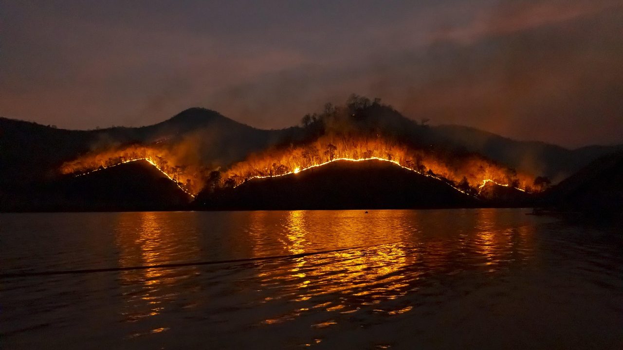 Thick orange wildfire smoke rises above a dry mountain ridge under a hazy sky.
