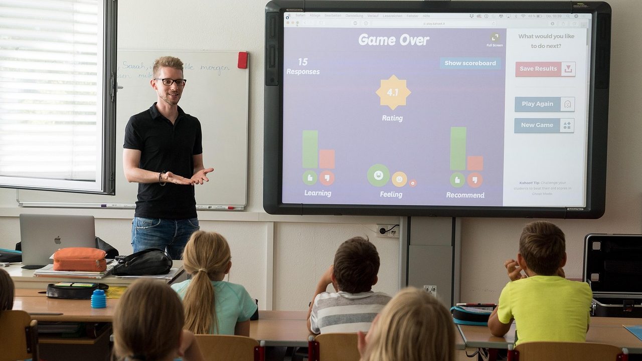 A teacher stands at a whiteboard in front of rows of students in a bright classroom.