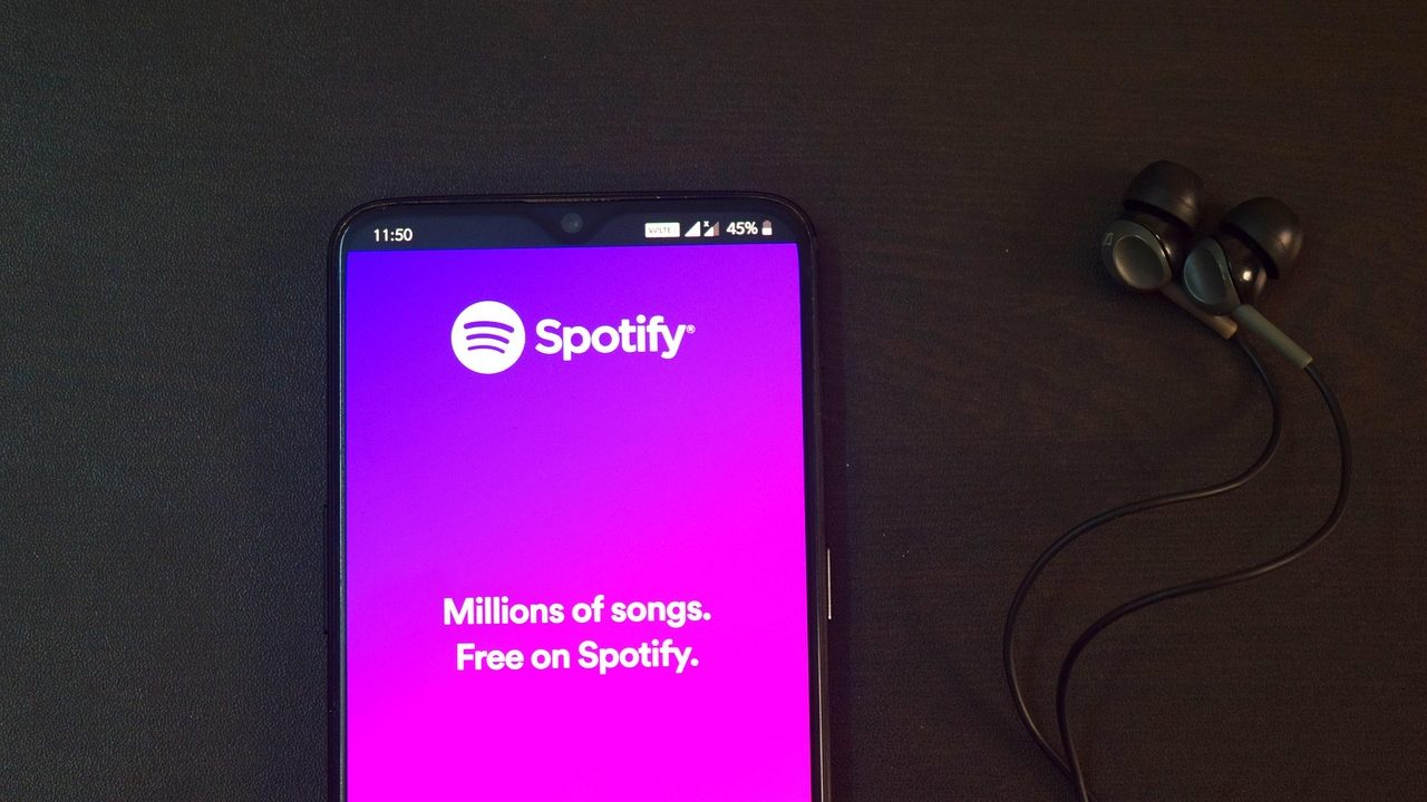 A smartphone displaying a music streaming app resting next to wireless earbuds on a desk.