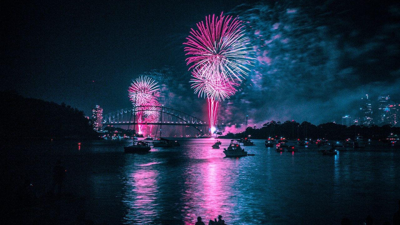 Colorful fireworks burst above a large suspension bridge reflected in dark water below.