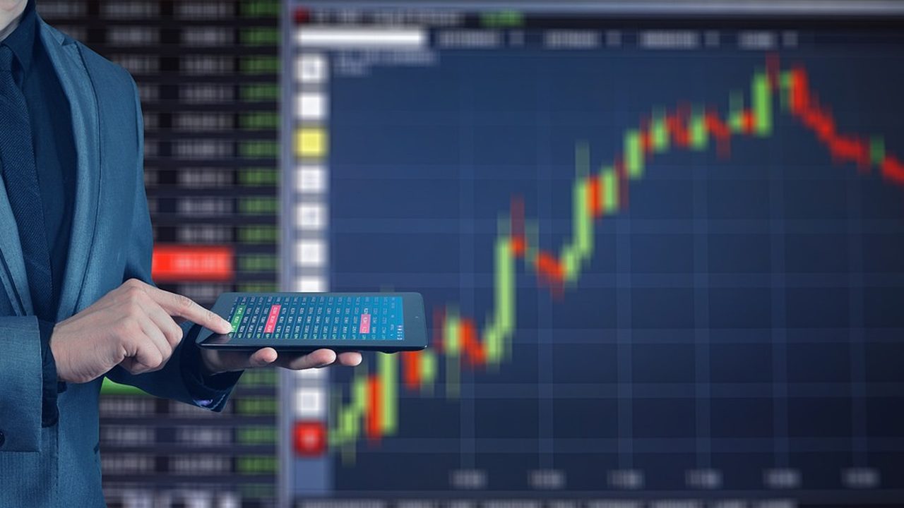 Traders stand at workstations on a busy stock exchange floor surrounded by multiple display screens.