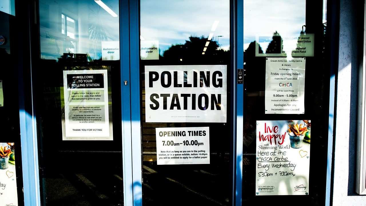 A row of blue voting booths inside a public polling station with American flags visible.