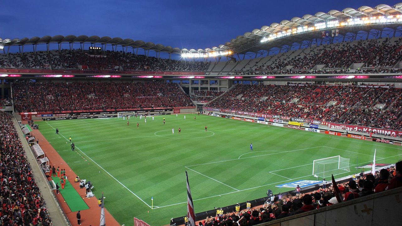 A packed soccer stadium at night with floodlights illuminating a green pitch.