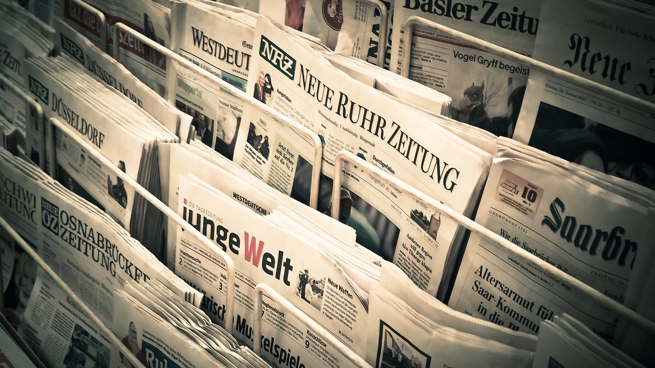 A stack of morning newspapers spread across a table with bold headlines visible.