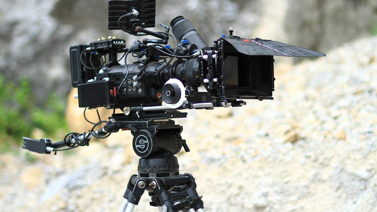 A film camera on a tripod stands on an empty movie set under bright studio lights.