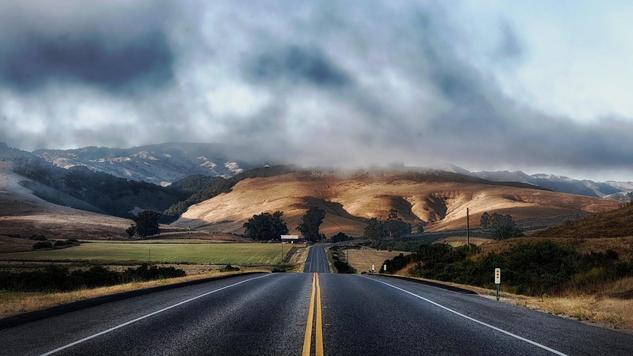 An empty two-lane highway cutting through dry hills under a partly cloudy sky.