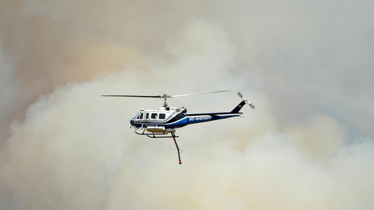 A plume of dark smoke rises above a dry canyon landscape under a hazy blue sky.