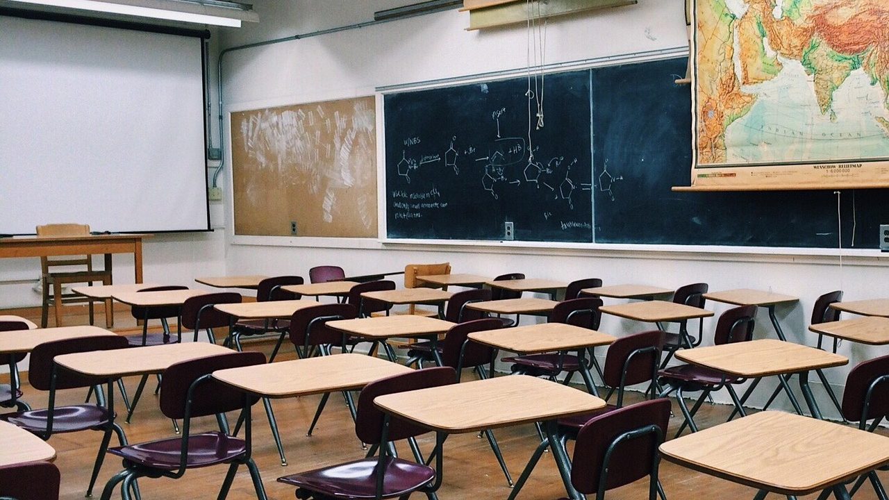 A vacant, sunlit classroom with small chairs, tables, and colorful bins arranged along the walls.