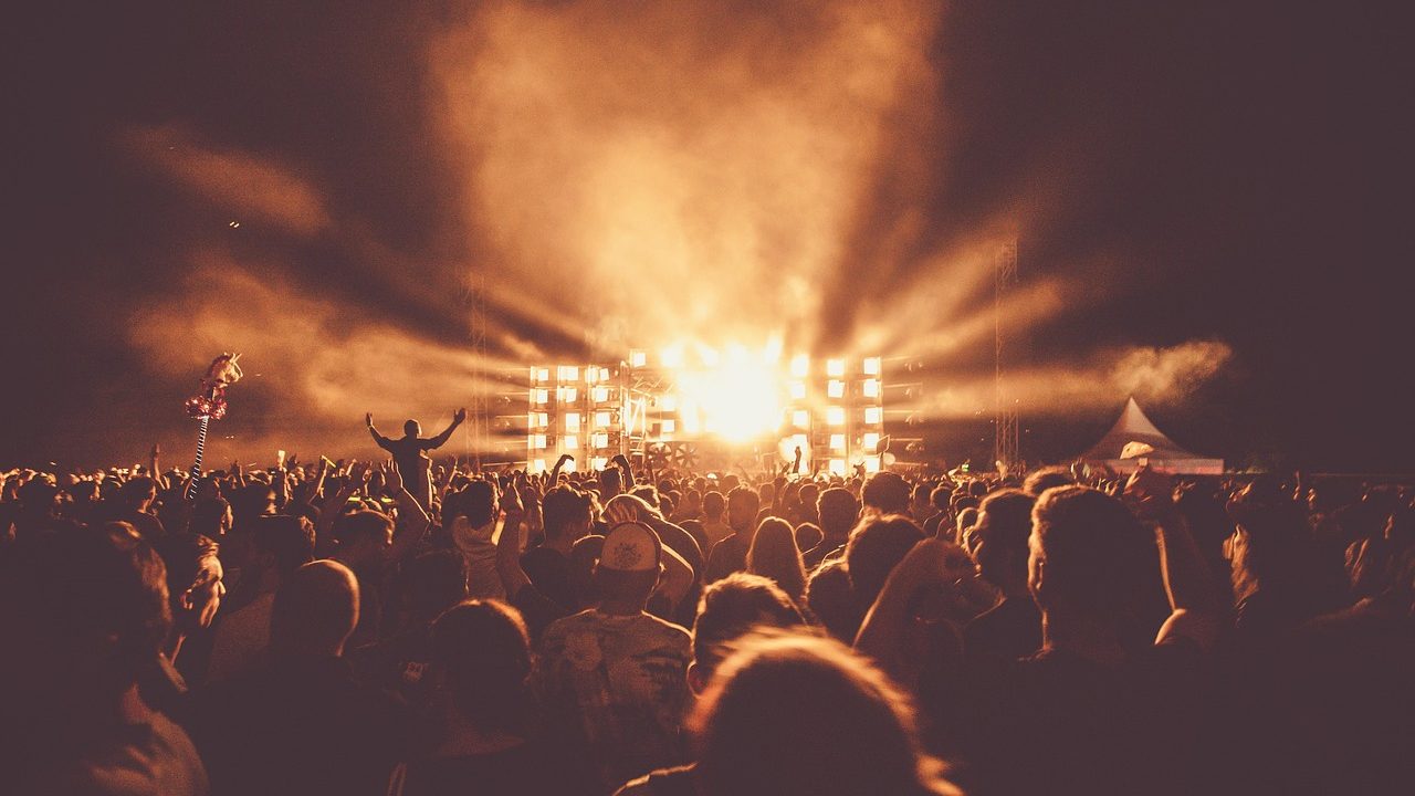 An open-air amphitheater glowing with stage lights as an audience fills the lawn at dusk.